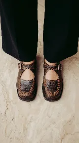 Mocasín de cuero marrón con acabado texturizado, diseño tipo Mary Jane con tira ajustable mediante hebilla lateral y detalle de costuras artesanales visibles en la puntera.