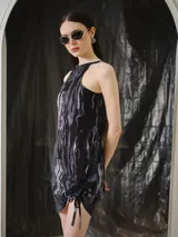 Vestido corto con escote alto y diseño sin mangas, confeccionado en tela liviana con estampa digital abstracta en tonos negros y grises. Presenta un frunce regulable con cordón en el lateral delantero que permite ajustar el largo.
