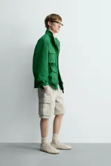 Chaqueta tipo cazadora de corte relajado en color verde, confeccionada en tejido de mezcla de algodón. Presenta cuello subido con forro interior a cuadros, manga larga con puños abotonados y cuatro bolsillos frontales de plastrón con solapa. Incluye cierre frontal de cremallera oculta por solapa con botones a presión y cintura ajustable en el interior.