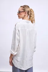 Camisa blanca de lino con cuello clásico, cierre frontal con botones y mangas largas con puños abotonados. Presenta apliques de encaje en hombros y bolsillos delanteros.