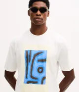 Remera de corte boxy confeccionada en algodón, con cuello redondo y manga corta. Presenta una estampa frontal con diseño artístico abstracto en tonos azules y marrones sobre un fondo claro.