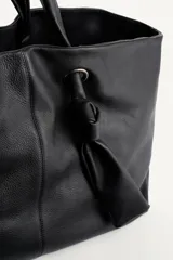 Bolso tipo shopper de cuero negro con textura granulada. Presenta doble asa de mano, cierre magnético y un detalle decorativo de nudo con argolla metálica en el frente.
