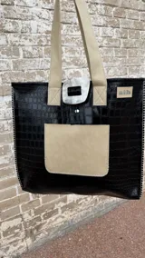 Tote bag de ecocuero negro con textura croco, cuenta con un bolsillo frontal de gamuza marrón y un detalle decorativo circular de guampa en el cierre superior. Posee doble asa de hombro y costuras visibles en los bordes.