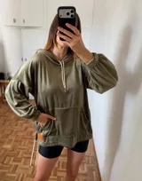 Buzo canguro con capucha, color verde oliva, confeccionado en tejido de terciopelo o velour. Presenta cordones blancos en la capucha y puños elásticos en mangas y ruedo. Posee un bolsillo canguro frontal.