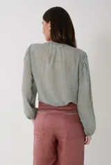 Camisa de voile de algodón color verde gastado con escote en V y aplique de guarda bordada en punto smock tostado. Presenta mangas largas abullonadas con puños elásticos y cierre frontal con pequeños botones de nácar.