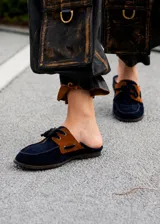 Mules estilo mocasín confeccionados en gamuza azul marino con detalles en gamuza color suela en los laterales. Presentan cordones frontales decorativos y una silueta abierta en el talón.