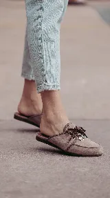 Zueco de cuero color marrón topo, destalonado, con cordones marrones y dijes metálicos colgantes. Presenta costuras artesanales a contratono y suela plana.