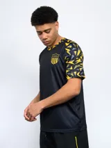 Remera deportiva de hombre, color negro, con cuello redondo y mangas cortas con estampado geométrico en amarillo y gris. Lleva el escudo de Peñarol en el pecho.