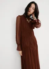 Vestido midi marrón de viscosa transparente, con diseño plisado, cuello alto fruncido y mangas largas abullonadas con puños elásticos. Presenta detalles de costuras fruncidas en la cintura y falda escalonada con vuelo.