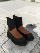 Botín de caña media con diseño bicolor, combinando cuero texturizado en color suela en la parte frontal y cuero liso negro en la puntera y talón. Presenta una caña de neopreno elastizado en color negro y suela de goma track con plataforma.