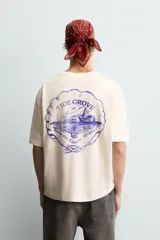 Camiseta de corte boxy fit, color beige con efecto lavado, cuello redondo y manga corta. Presenta un estampado gráfico en azul oscuro en el frente con tipografía estilo vintage.