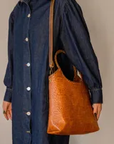 Bolso tipo matera de cuero con textura de reptil, color tostado, con manijas trenzadas y detalles de costura artesanal en los bordes.