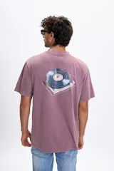 Remera de manga corta con corte oversize, color violeta con efecto lavado. Presenta una estampa gráfica en la espalda que ilustra un disco de vinilo sobre una bandeja giradiscos con texto decorativo.