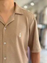 Camisa de manga corta color blanco marfil, confeccionada en tejido texturizado con cuello camisero, cierre frontal de botones y un pequeño bordado oscuro en el pecho.