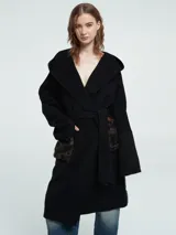 Abrigo largo de paño negro con capucha, cierre cruzado con lazo ajustable en la cintura y bolsillos delanteros con textura de pelo sintético. Presenta un diseño con espalda en nylon capitoneado.