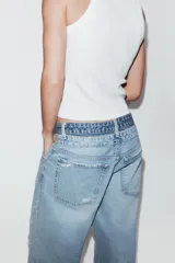 Jeans de tiro medio, color azul claro, con doble cinturilla y trabillas. Presenta cinco bolsillos, detalles de rotos y cierre frontal con cremallera y botones.