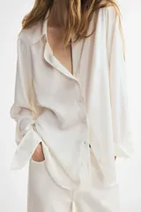 Blusa blanca de tela con brillo, cuello clásico, escote en V con botones frontales, mangas largas con puños sencillos y bajo redondeado.