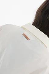 Camisa blanca de mangas largas con cuello clásico y cierre con botones. Presenta detalles de fruncido vertical en la parte delantera y cordones ajustables en los laterales. Corte holgado.