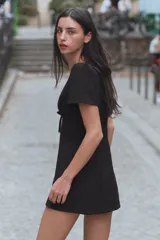 Vestido mini negro con escote en V amplio, mangas cortas abullonadas y detalle de lazo en el delantero. Cuenta con cierre de cremallera oculta en la espalda.