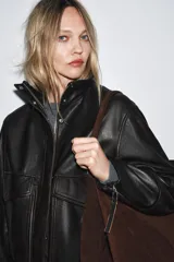 Chaqueta bomber de cuero negro con cuello alto, manga larga con cremalleras metálicas, bolsillos de plastrón con solapa y botón en delantero, bajo acabado en elástico y cierre frontal con cremallera y botones a presión ocultos por solapa.