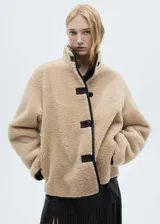 Chaqueta de efecto pelo en color beige, con diseño oversize y corte recto. Presenta cuello alto, cierre frontal con botones y detalles de ribetes en cuero sintético negro.