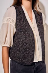 Chaleco sin mangas de corte al cuerpo, color negro con bordados florales en hilo contrastante. Presenta un diseño abierto con terminaciones reforzadas.