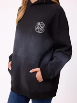 Canguro oversize color negro con efecto degradado, capucha, bolsillo frontal tipo canguro y puños elásticos. Presenta un parche con logo en la manga.