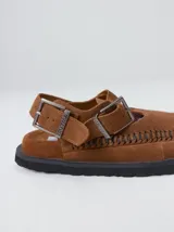 Slipper tipo guillermina confeccionada en gamuza color marrón, con plataforma de goma negra y doble hebilla ajustable. Presenta un pespunte decorativo en el borde inferior y cuenta con interior recubierto en cuero.