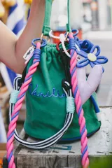 Cartera tipo bucket verde con base estructurada, cordón de ajuste rosa y azul, y llaveros colgantes de flor azul y corazón rosa.