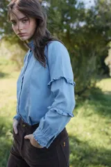 Camisa de jean azul claro con cuello alto, cierre frontal de botones y mangas largas con detalles de volados horizontales y puños con volados.