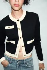 Chaqueta de punto de corte recto y manga larga, con cuello redondo. Presenta un diseño bicolor en negro y crema, con cierre frontal de botones dorados y bolsillos de plastrón en el delantero con detalles en contraste.
