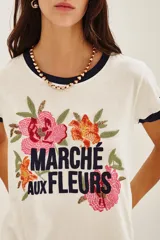 Remera de algodón color crema con cuello y bordes de mangas en contraste azul marino. Presenta un diseño frontal con bordados de flores coloridas y la inscripción 'MARCHÉ aux FLEURS' en letras azules.