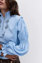 Blusa celeste de manga larga con puños fruncidos y elásticos, escote en V con cuello alto con volados y delicados bordados florales en el pecho.