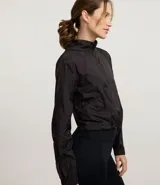 Campera cortavientos deportiva de corte cropped, color verde oscuro. Presenta capucha, cierre frontal con cremallera, mangas largas con puños elásticos y cordón ajustable en el ruedo.