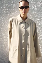 Trench largo de Zara Woman Collection, confeccionado en color beige. Cuenta con cuello de solapa, manga larga, bolsillos frontales de parche y cierre central con botones visibles.
