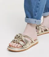 Sandalia plana de color negro con diseño de tiras tubulares acolchadas tipo puffer entrelazadas con nudos. Presenta puntera redonda, plantilla anatómica y suela de goma al tono.