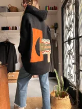 Campera de paño color negro con capucha y diseño de collage en la espalda. El collage incorpora parches en tonos naranja, verde oliva y un tejido con patrón geométrico étnico.
