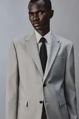 Blazer slim fit de color gris, con cuello de solapas de muesca, cierre frontal de dos botones, bolsillos laterales con solapa y bolsillo de vivo en el pecho. Presenta mangas largas con botones en los puños y aberturas en la espalda.