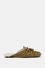 Mocasín destalonado tipo mule confeccionado en serraje color tostado, con detalle de fruncido y borlón en la parte delantera. Interior forrado en efecto borreguito y suela plana.