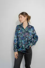 Camisa de seda con cuello clásico y cierre frontal de botones, de manga larga con puños abotonados. Presenta un estampado floral en tonos azules, verdes y rosados sobre fondo negro.