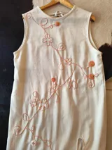 Vestido midi de lino de algodón color crudo, sin mangas, con escote redondo y delicado bordado a mano en hilo color naranja con motivos florales y vegetales en diagonal.