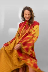 Vestido largo plisado con estampado abstracto en tonos rojo, naranja y dorado, cuello alto y mangas acampanadas. Incluye lazo fino para ajustar en la cintura.