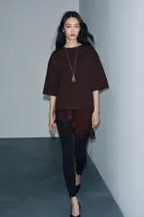 Camiseta de algodón de corte regular, color marrón oscuro o negro, con cuello redondo y manga corta. Presenta un bajo asimétrico con aplicación de encaje negro a tono.