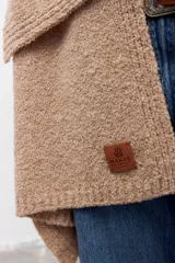 Chal de tejido de punto en color beige, con textura suave y diseño envolvente. Presenta una etiqueta de cuero sintético con logo grabado en la parte inferior.