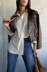 Camisa beige de corte oversize, con cuello clásico y cierre frontal de botones. Presenta un diseño bordado detallado en la parte superior delantera, con mangas largas y un estilo versátil.