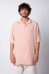 Camisa de manga corta con cuello cubano, confeccionada en fibrana premium. Presenta un corte oversize, cierre frontal con botones y un diseño minimalista en color rosado.