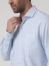 Camisa de manga larga de algodón celeste con sutil patrón a cuadros, cuello camisero, cierre frontal con botones y un bolsillo en el pecho con logo bordado.