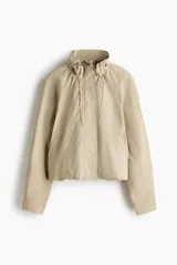 Campera corta de sarga de algodón, color beige, con cuello elevado ajustable por cordón, solapa rompevientos con botones a presión ocultos y cierre. Presenta bolsillos delanteros insertados, mangas ranglán con hombros caídos y bajo globo que genera una silueta abullonada.