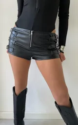 Short de cuero sintético negro con cierre frontal y detalles de tiras con hebillas metálicas en los laterales.