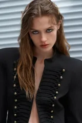 Blazer corta de estilo militar en color negro, con cuello redondo, manga larga y hombreras marcadas. Presenta un diseño frontal con bordados decorativos a tono y botones dorados, cierre con gancho metálico y forro interior.